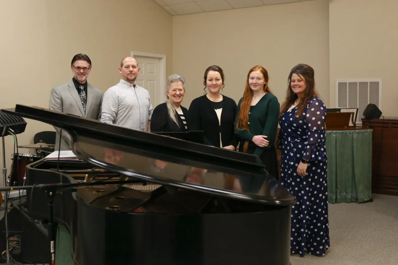 Group standing behind a grand piano indoors.