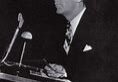 Man speaking at a podium, black-and-white photo.